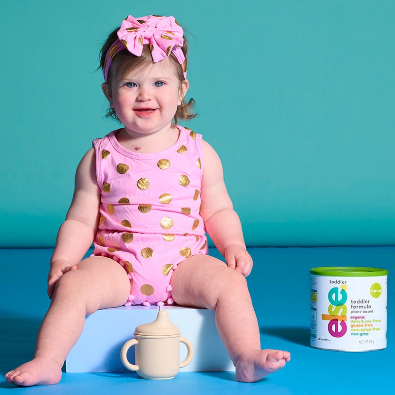 A baby sits on a blue box, holding a container of baby food, with a curious expression.