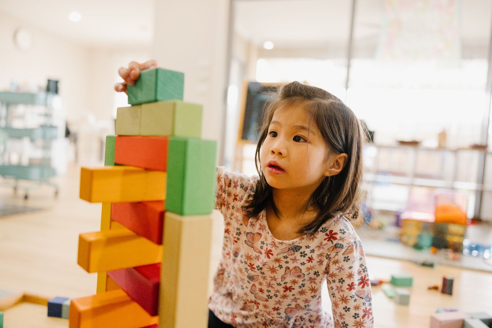 A preschooler engages in construction play, carefully stacking blocks both horizontally and vertically to form a tower, with warm-toned blocks at the bottom and green hues on top."