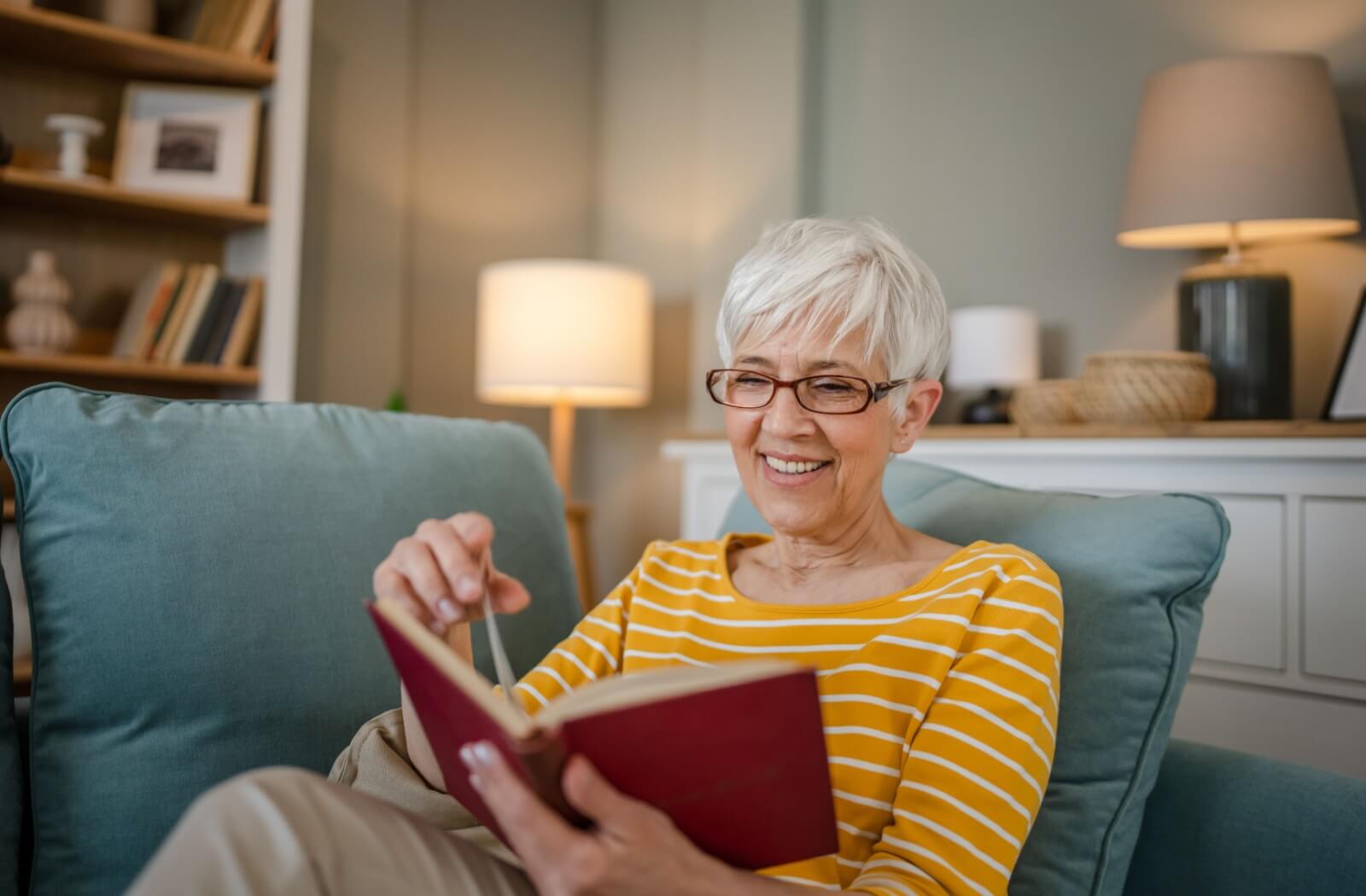 A smiling senior enjoys reading a book in preparation for the weekly book club discussion.