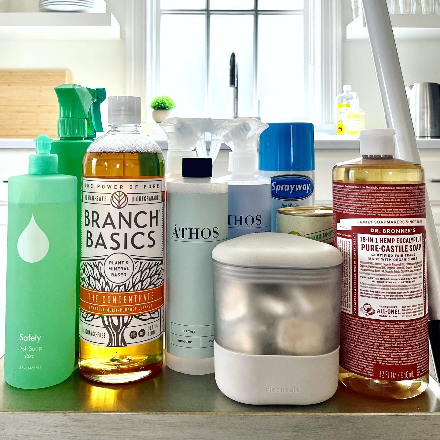 A variety of cleaning products lined up on a kitchen counter, including spray bottles, soap dispensers, and a cleaning tablet container. 