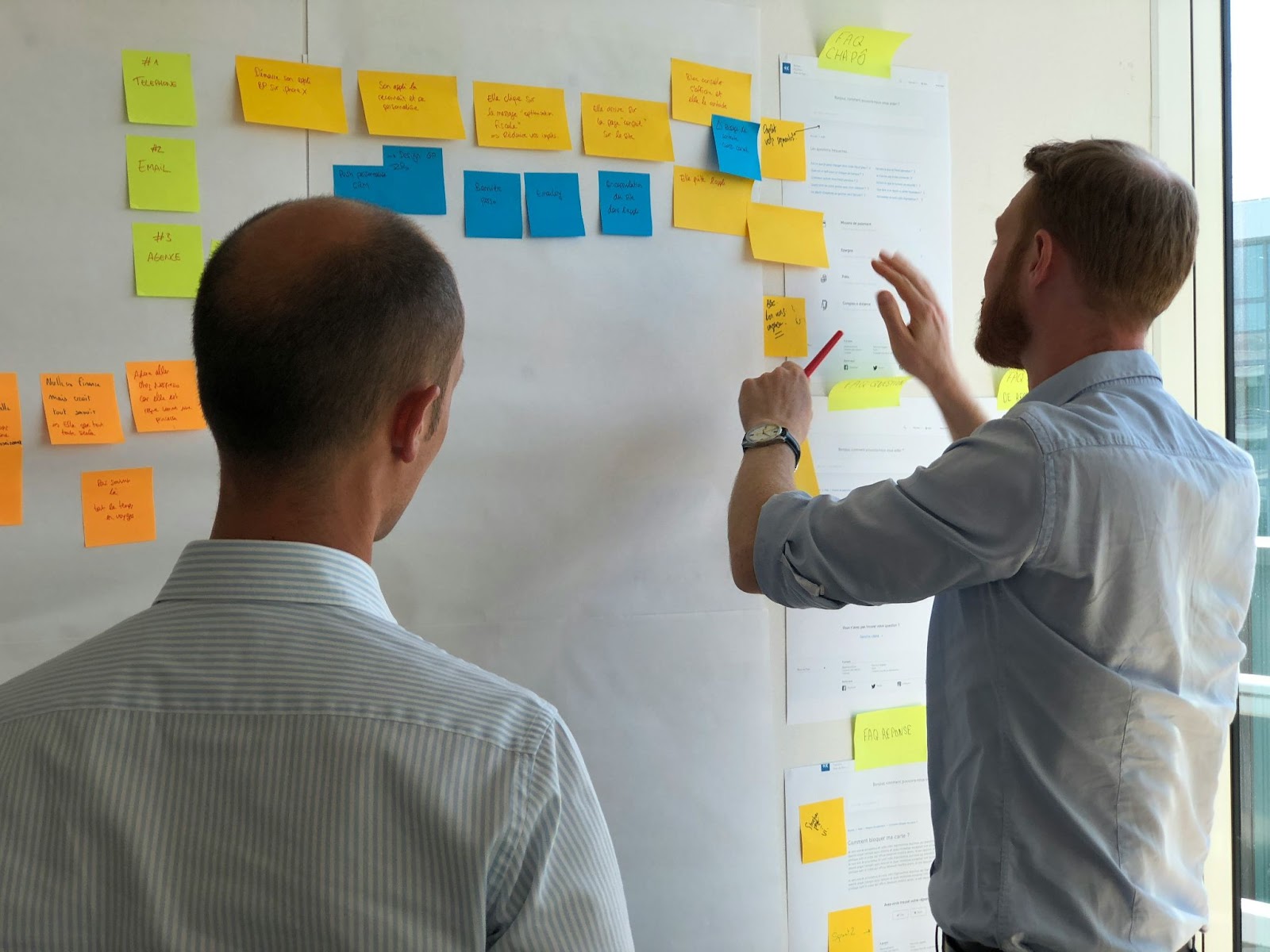 Two business professionals are standing in front of a whiteboard covered with colorful sticky notes, discussing and organizing tasks or strategies.