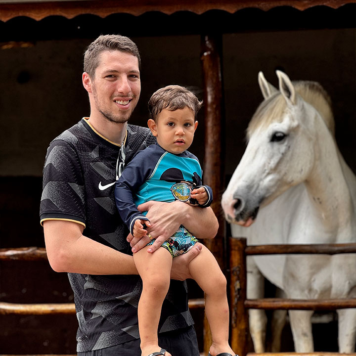Homem com menino no colo, ambos sorrindo, ao lado de um cavalo branco, na fazenda. Uma cena de alegria e conexão com animais, destaque para passeio a cavalo.