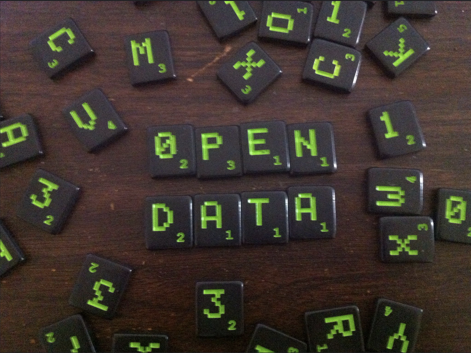 A photograph of a dark wooden tabletop with black tiles with bright green letters and numbers on them from the game Scrabble that are scattered across the table with the words “open data” made up of tiles in the middle of randomly arranged ones