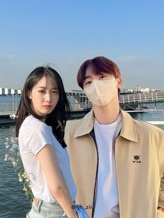 This contain SEVENTEEN Seungkwan and VIVIZ SinB standing next to each other near the water with a mask covering their mouths