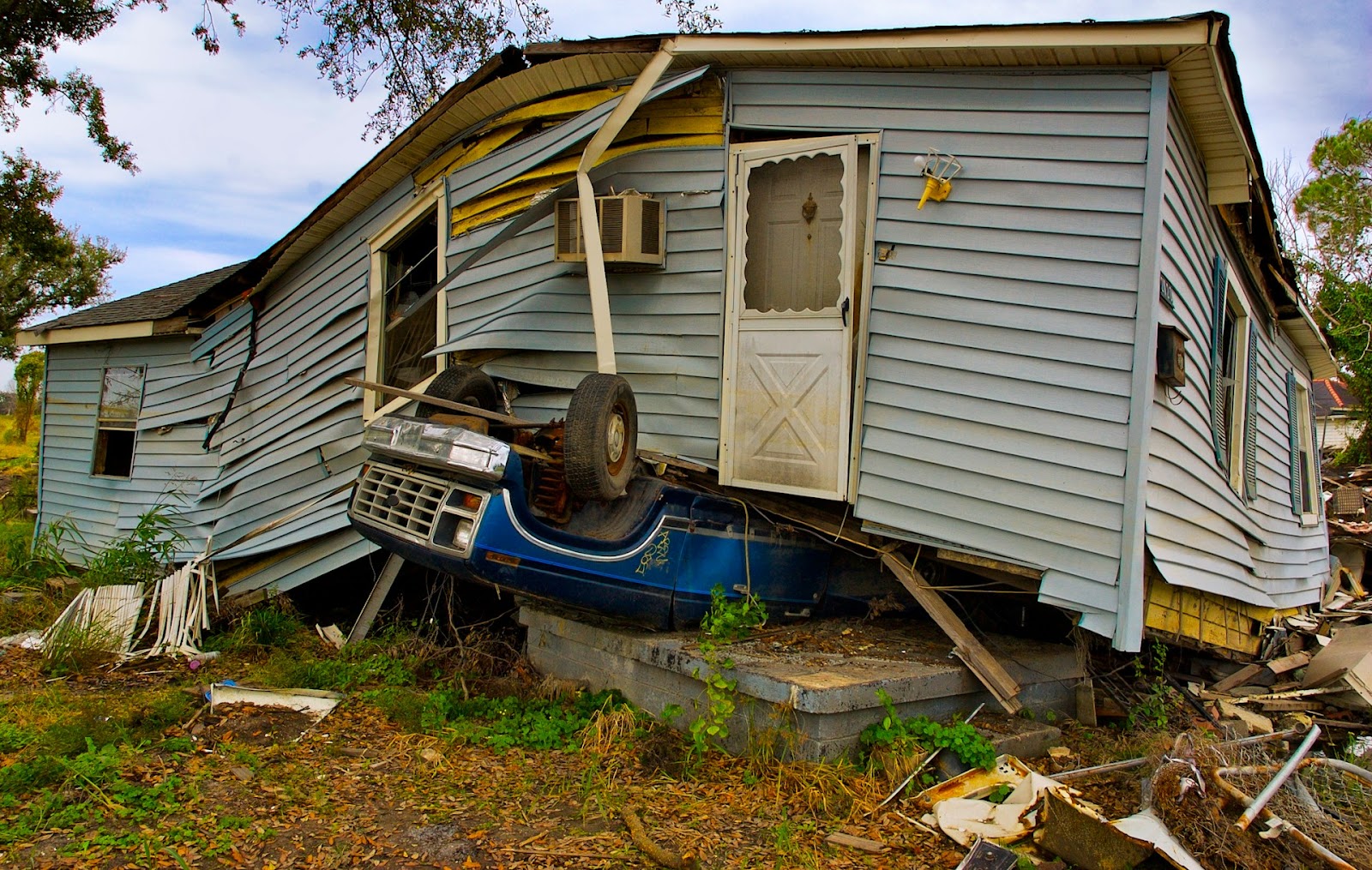 A gray house severely damaged and tilted with a blue truck crashed into its front corner.