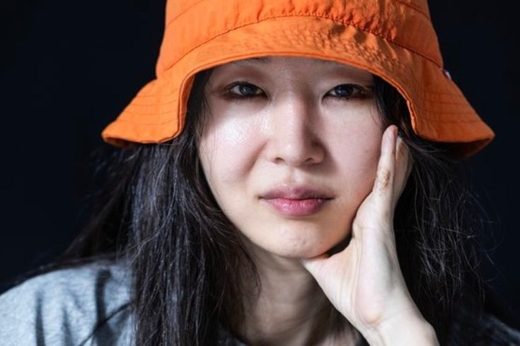 This contains an image of Min Hee Jin wearing an orange hat with her hand on her chin and looking at the camera