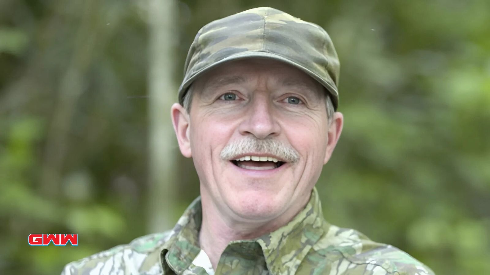 Mark Rogers in camouflage cap and jacket smiling in a forested background.