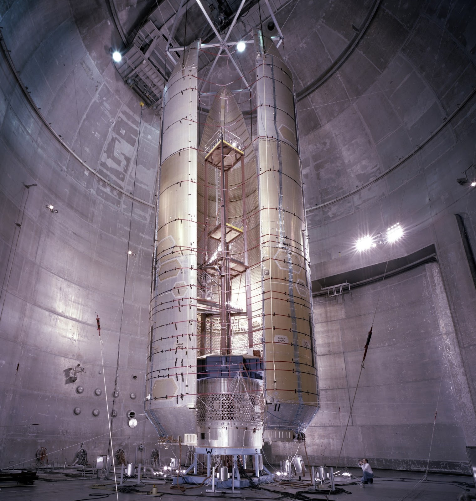 A very tall brown and silver spacecraft shroud stands inside a large grey chamber and towers over a person, seen at its bottom right.