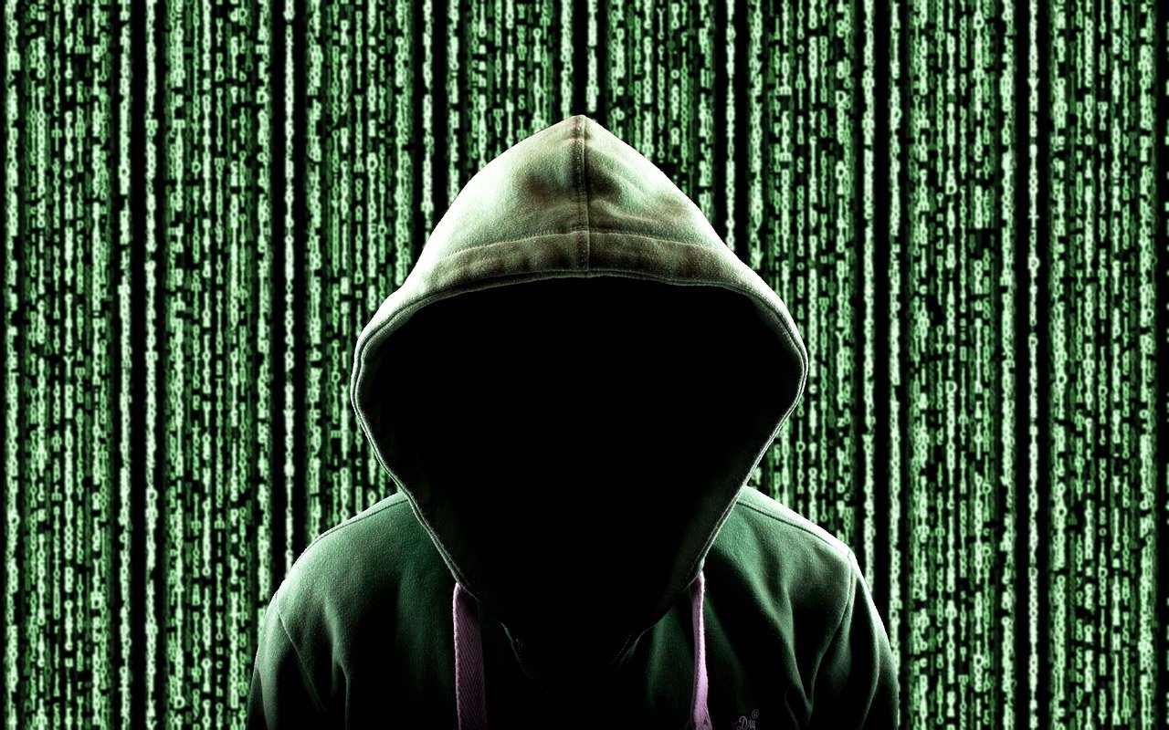 [hooded figure in front of a background of green binary code, evoking a hacker in cyber attack