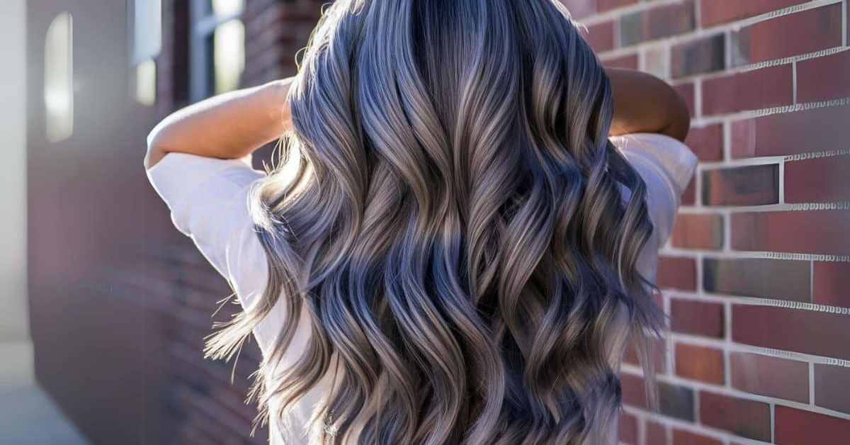 A woman with long, wavy hair stands before a brick wall, showcasing the beauty of Gray Blending for Natural Hair.