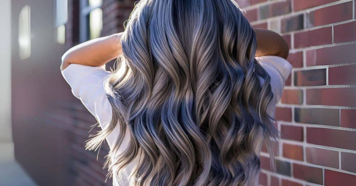 A woman with long, wavy hair stands before a brick wall, showcasing the beauty of Gray Blending for Natural Hair.