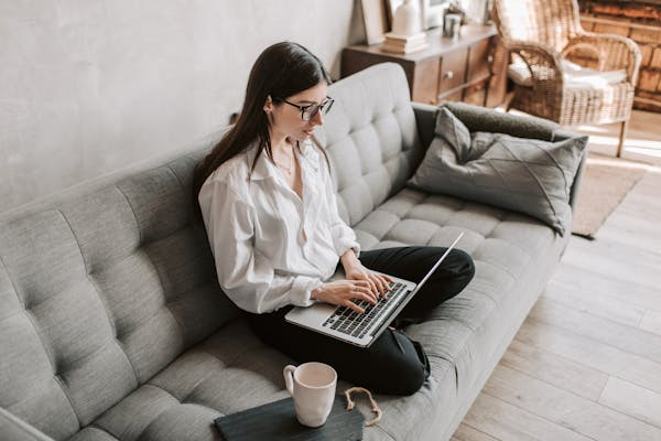 Top view of a young woman sitting folded legs at sofa, writing meeting minutes in a laptop, with a cup of coffee beside her