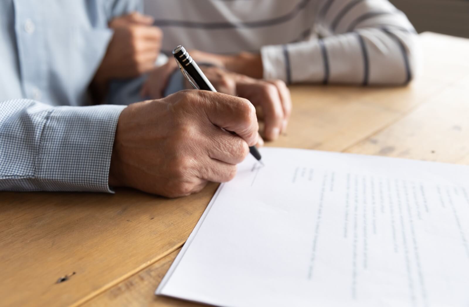 A close-up of an elderly person signing a document on a wooden table, with their elderly spouse in the background