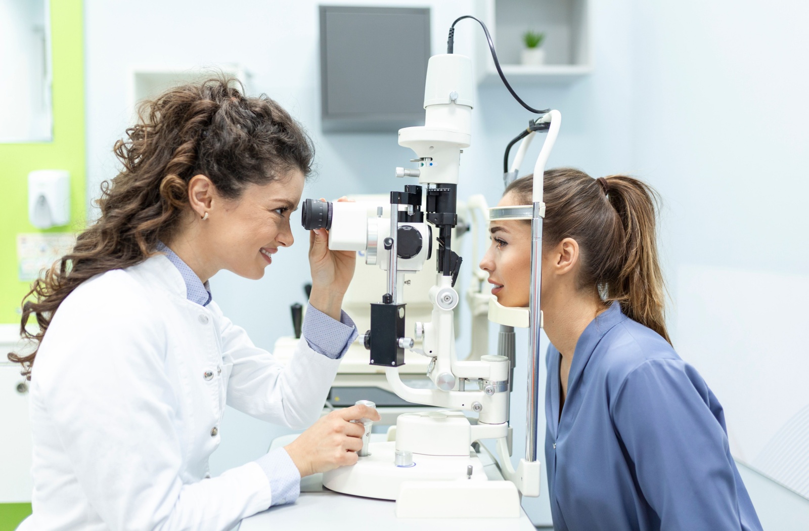 An optometrist smiling while examining a young pregnant woman's eyes to find out what's causing her eye twitching.