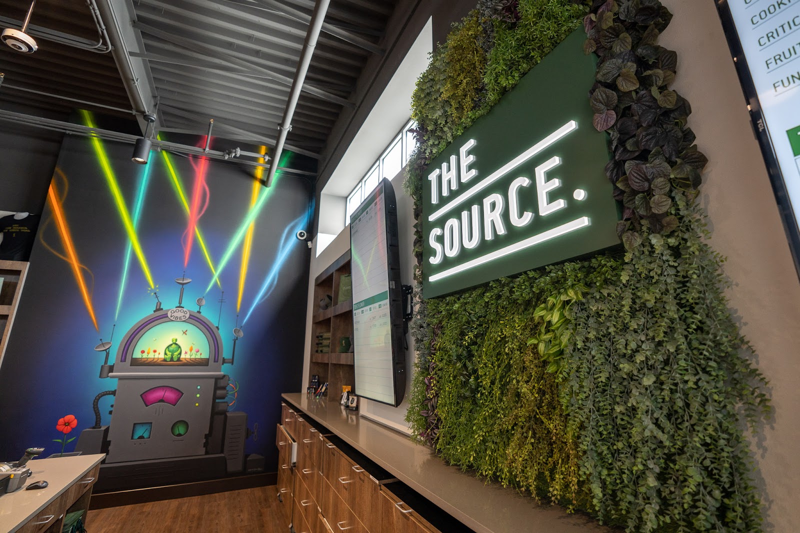 A lit up sign reads, The Source on a leafy green wall. On the opposing wall, a mural features a "Good Vibes Machine," with colorful laser beams pointing of it. 