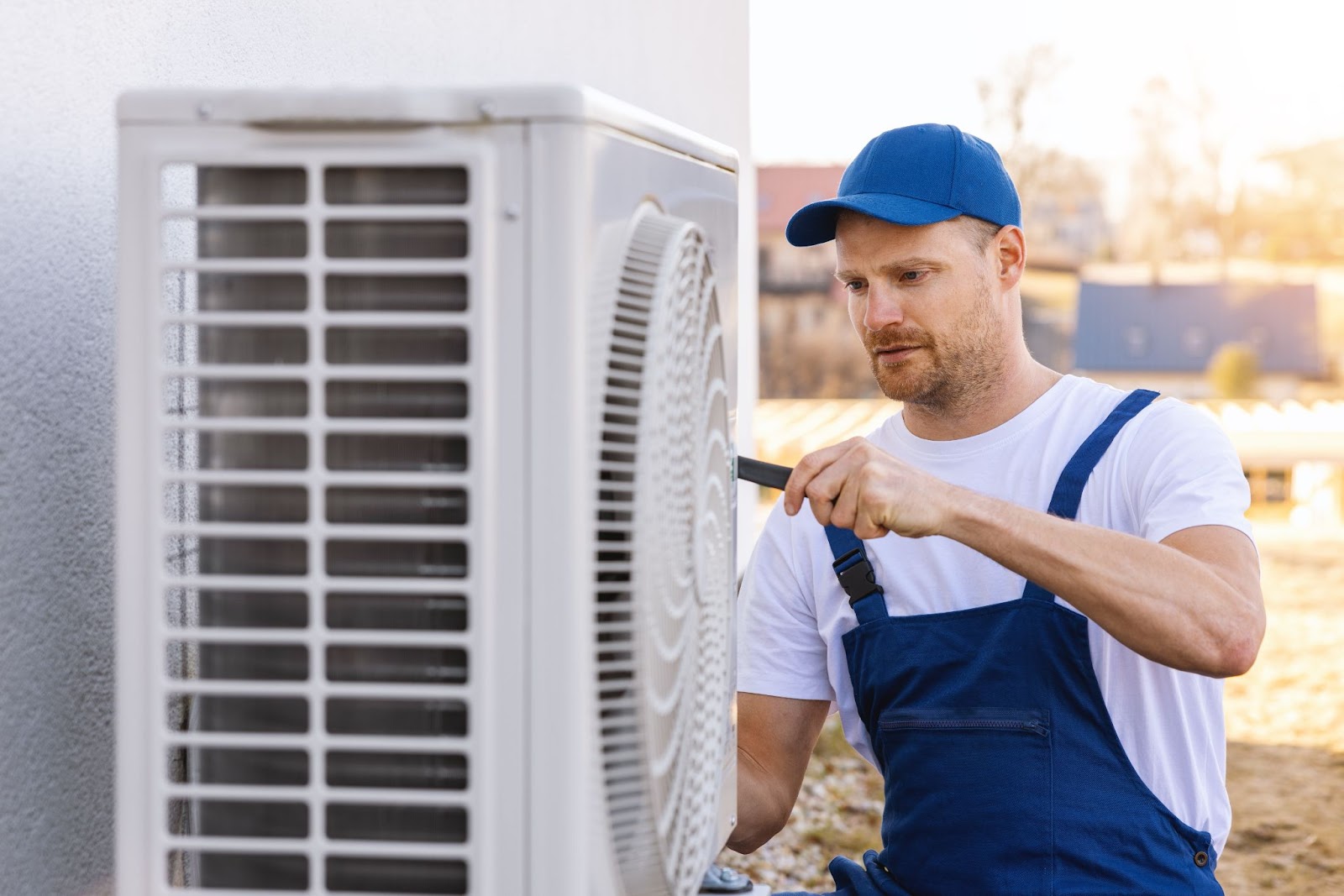 An HVAC technician wearing a blue hat and overalls while working outdoors on an outdoor HVAC unit