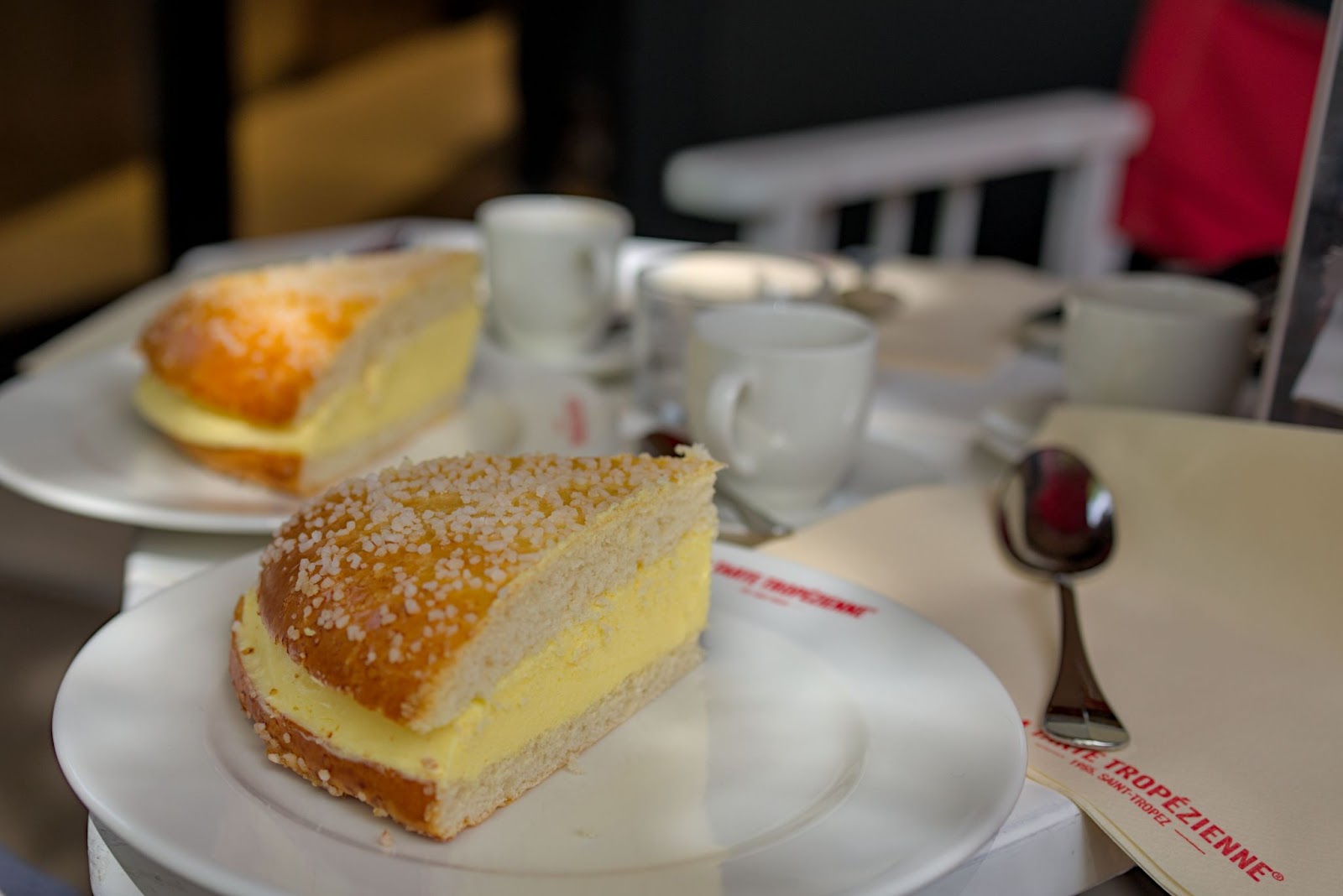 Close-up of two slices of Tarte Tropézienne, a cream-filled brioche dessert topped with pearl sugar, served on white plates with coffee cups and spoons at a café table in Saint-Tropez.