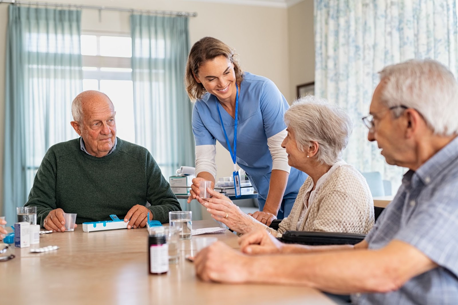  3 older adults sitting at a table as a smiling nurse helps them with medication.