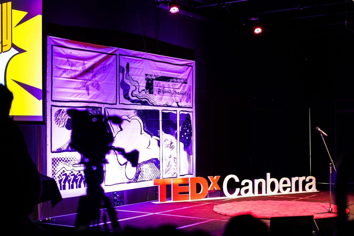 A camera on a tripod pointed at a stage with a sign reading 'TEDXCanberra' on it