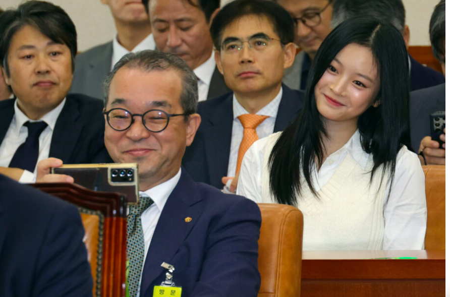 This is a picture of newjeans Hanni in white shirt sitting in the mist of men in suits and one of the man in front of her taking a selfie