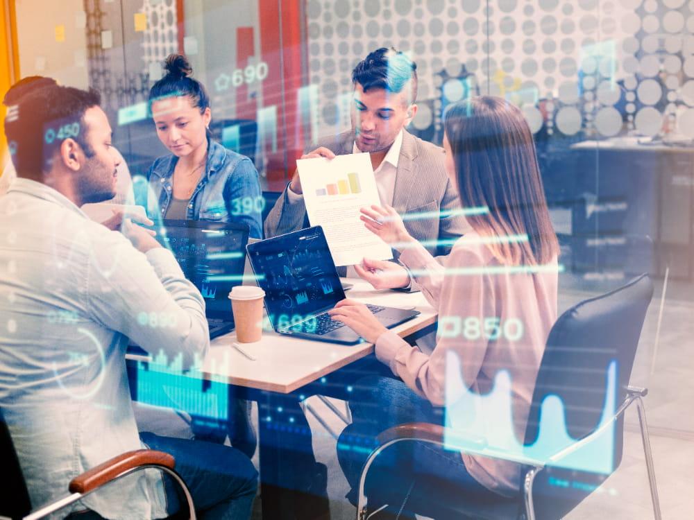 Profissionais participando de uma reunião em torno de uma mesa, utilizando laptops e analisando gráficos em uma apresentação.