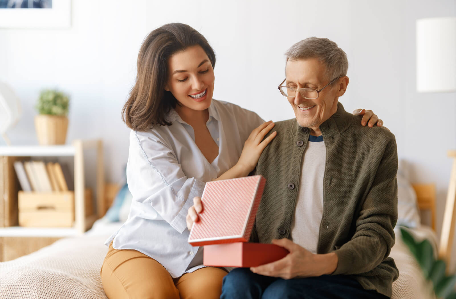 A senior sits on their bed beside their adult child, opening a gift. The senior's face is lit up with joy, and their child has their arms around their parent's shoulders in a loving manner.