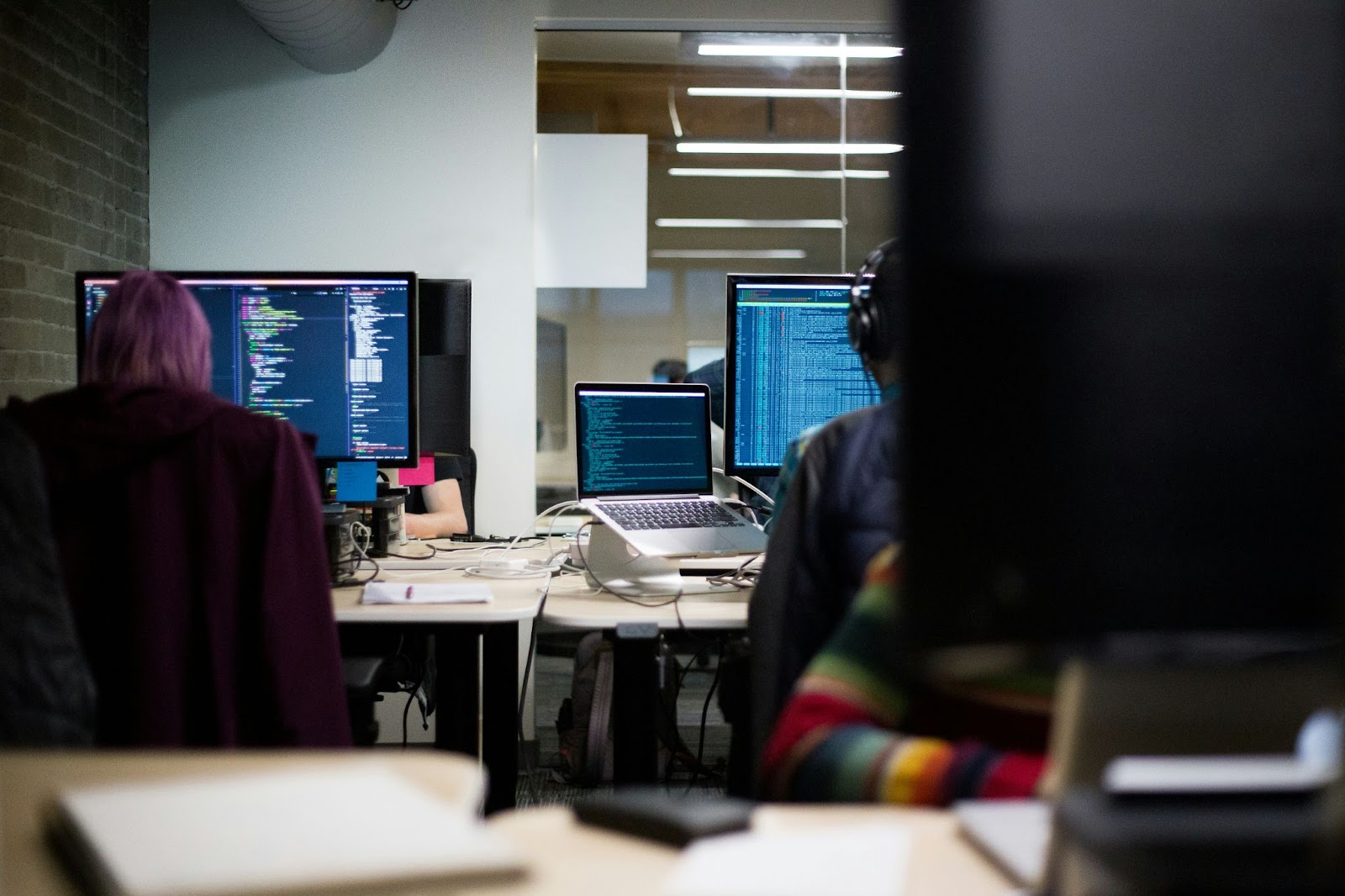 Two people work at a desk with multiple computer monitors displaying code and data in a modern office environment.