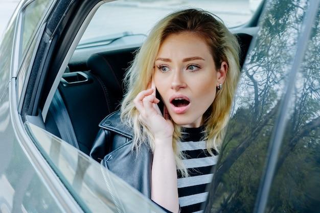 Young surprised woman talking on smartphone while travelling in the back seat of car wearing striped top and black leather jacket