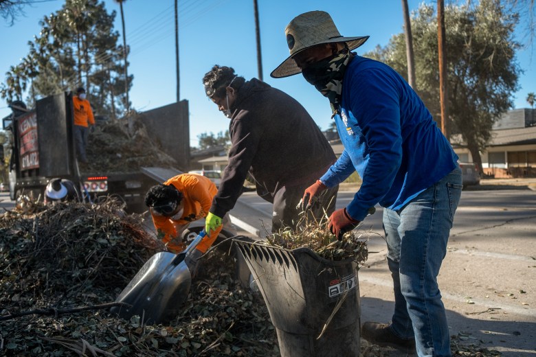 Workers clear debris into a trash bin on a residential street, with one person in a wide-brimmed hat and another in safety goggles. A truck filled with greenery is parked nearby under a clear blue sky, and trees line the background. The scene captures teamwork in an outdoor cleanup effort.