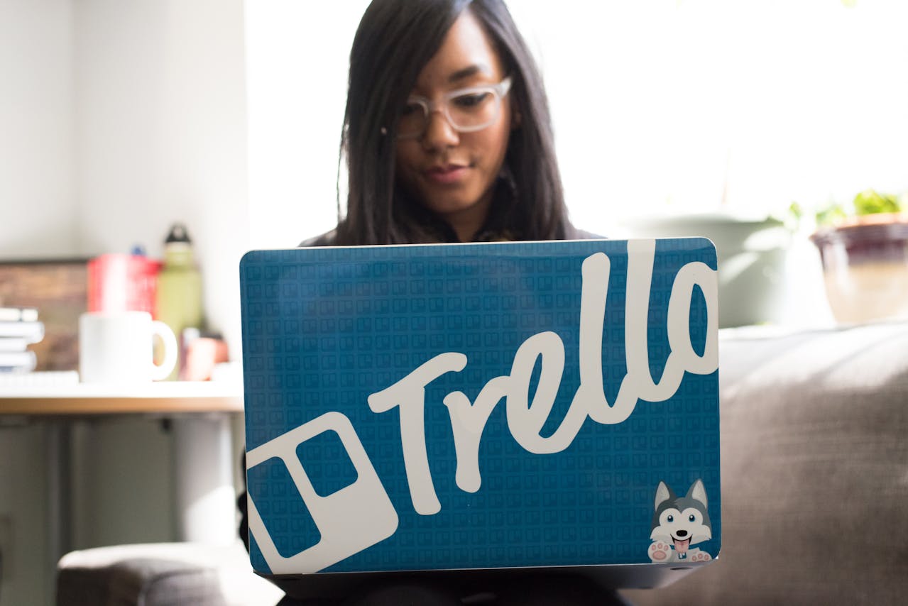 Woman using a laptop with a Trello sticker, working on project management tasks.