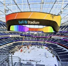 This contain  SoFi Stadium  filled with lots of seats and people standing around it in front of a colorful sign that reads soft stadium