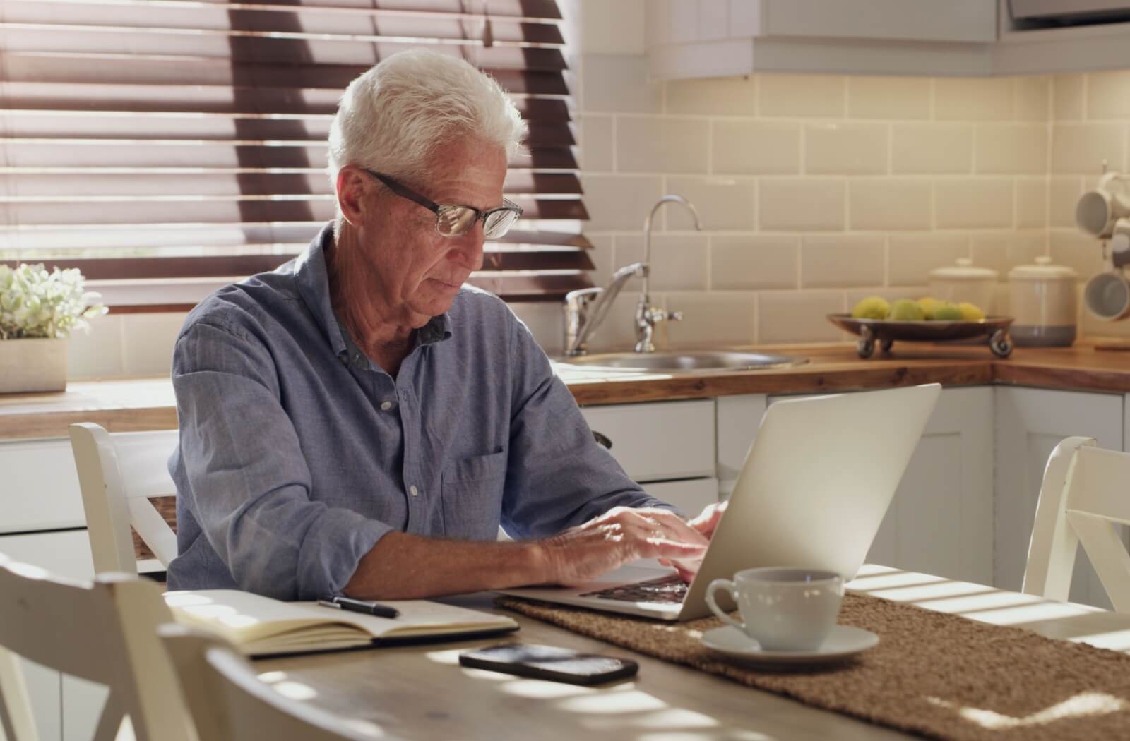  An older adult man sitting at his kitchen table, drinking tea while writing an email to a family member