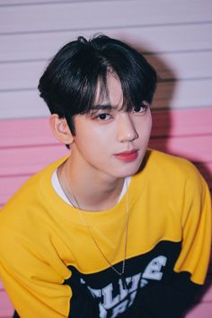 This contains an image of ZEROBASEONE's Yujin with a black hair and yellow shirt sitting in front of a pink wall