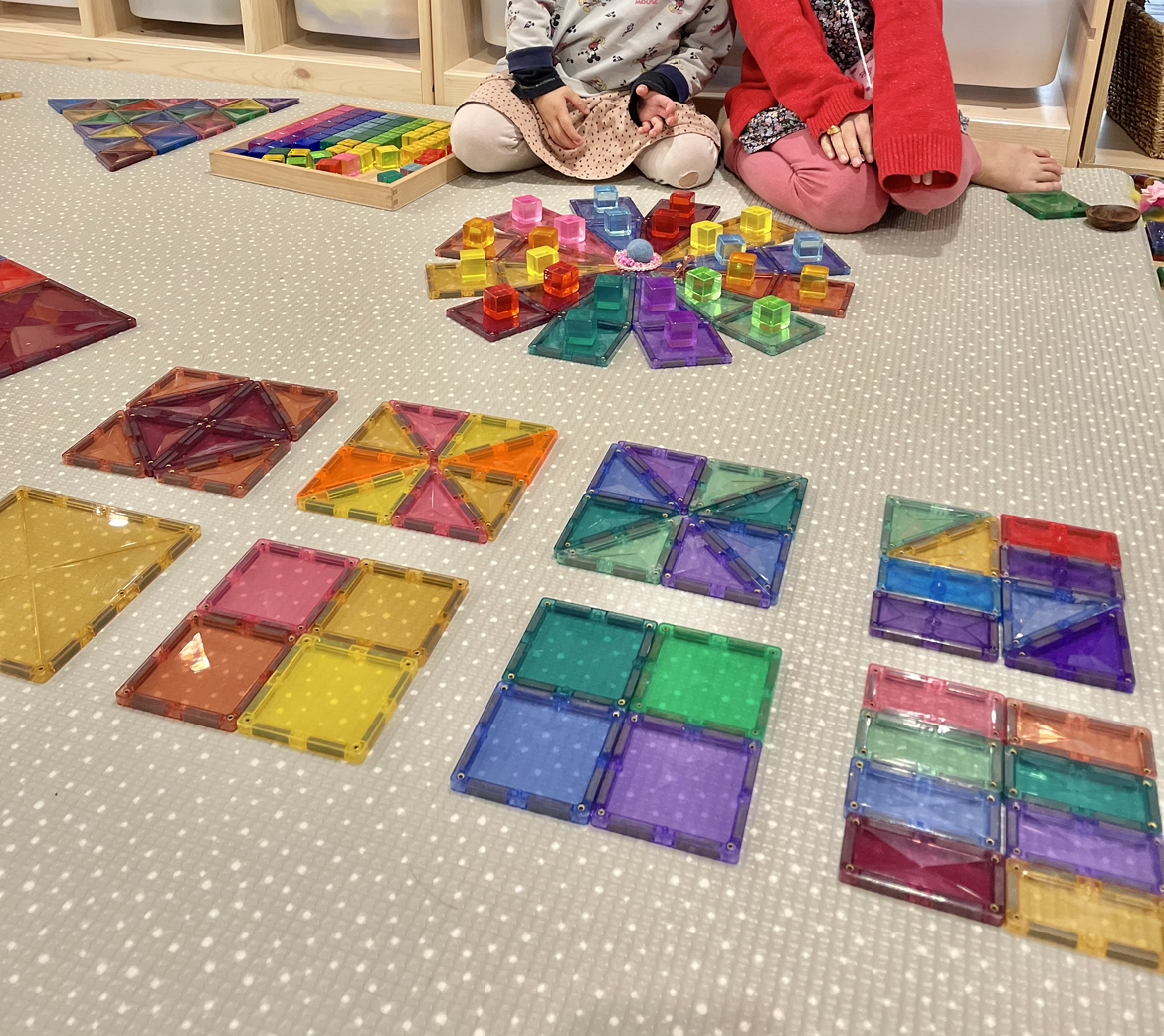 Children explore shapes, patterns, and colors by arranging four small squares and eight rectangles to create larger shapes, including cool and warm color tiles. They also construct a snowflake pattern with magnetic tiles and decorate it with rainbow-colored acrylic blocks, showcasing their creativity in combining different elements to form a cohesive design.
