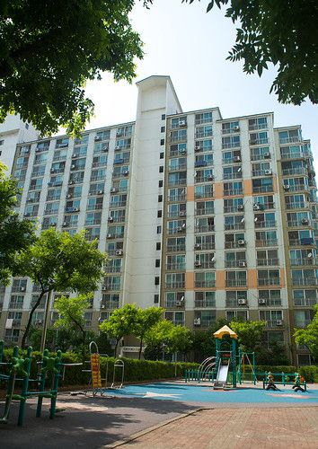 Foto bangunan apartemen bertingkat tinggi dengan banyak unit, dilengkapi area taman bermain anak dan alat kebugaran luar ruangan di bagian depan, dikelilingi pepohonan hijau.