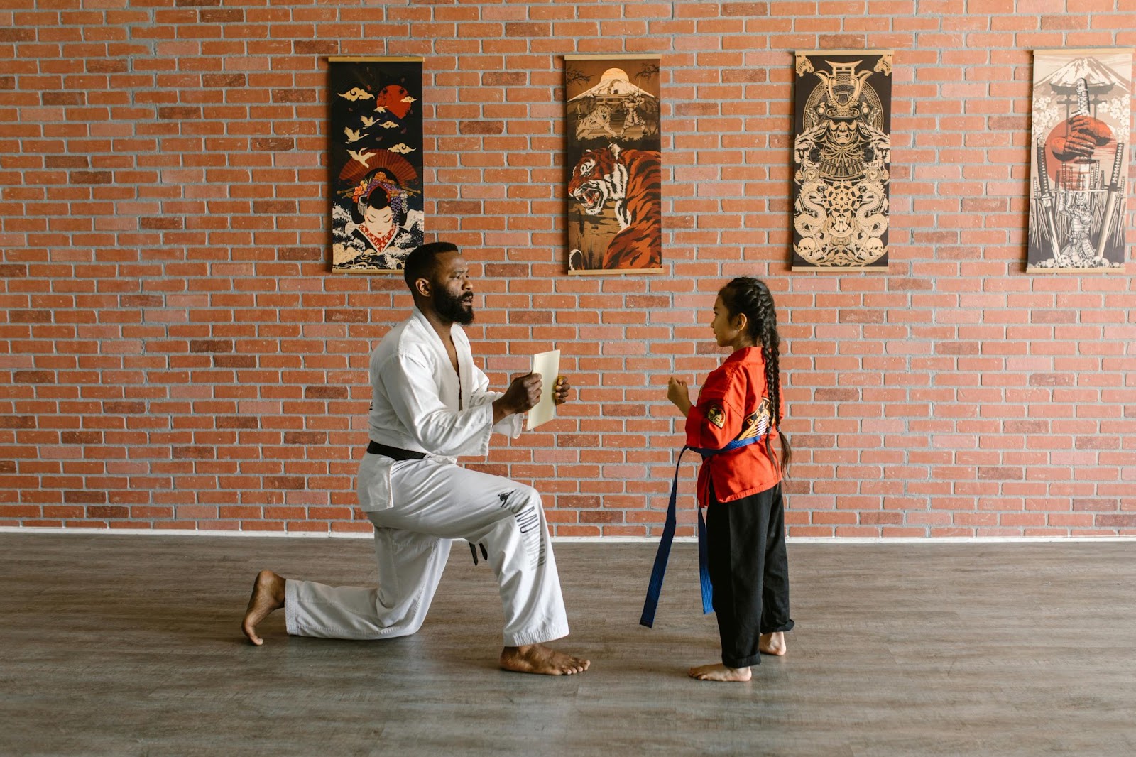 A martial arts instructor kneels on one knee, holding a piece of paper at arm's length in front of a young student.