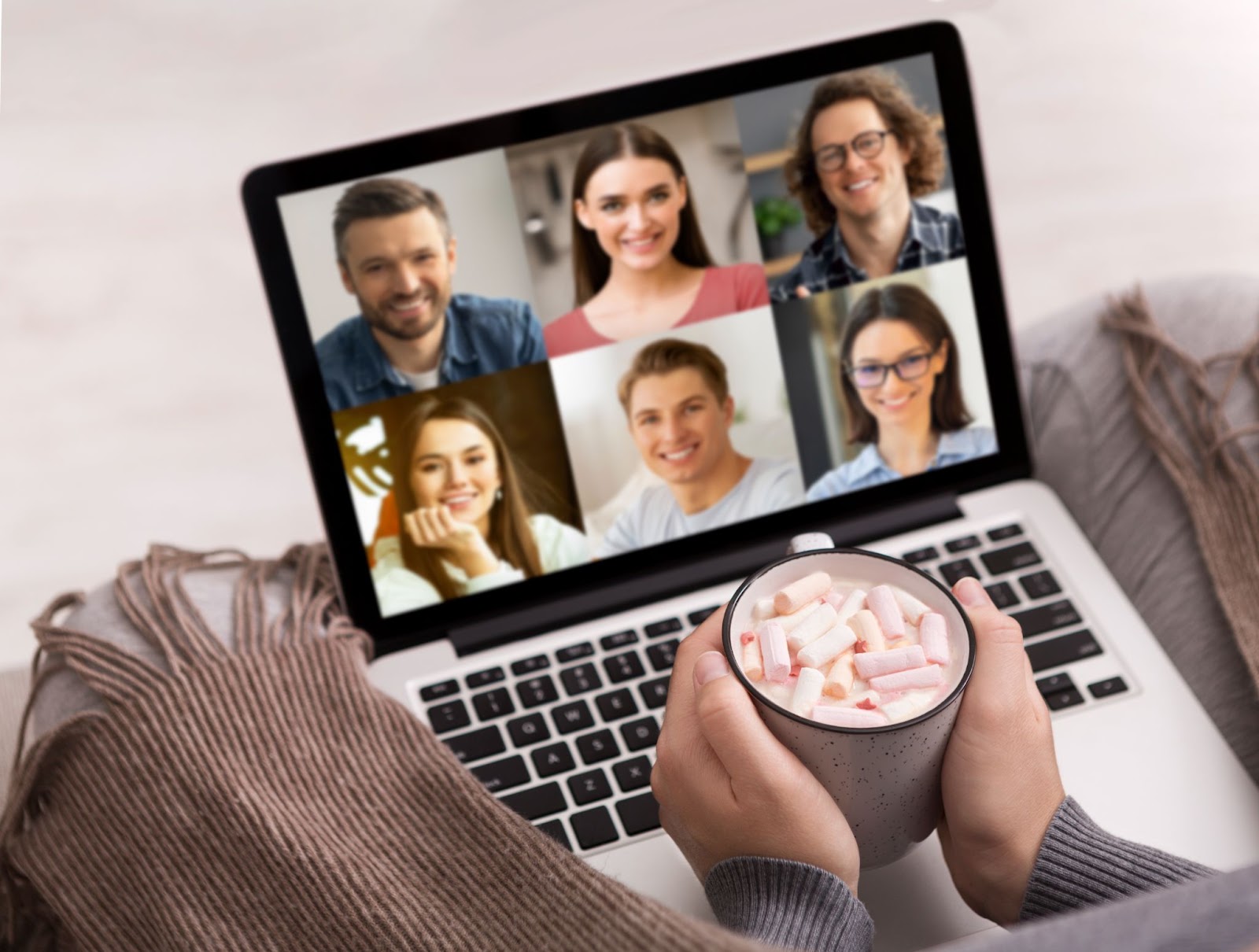 (Person holding a cup of hot chocolate topped with marshmallows while participating in a video call with six people on a laptop screen. A brown striped blanket is partially covering the laptop, making it feel like a cozy show and tell session.)