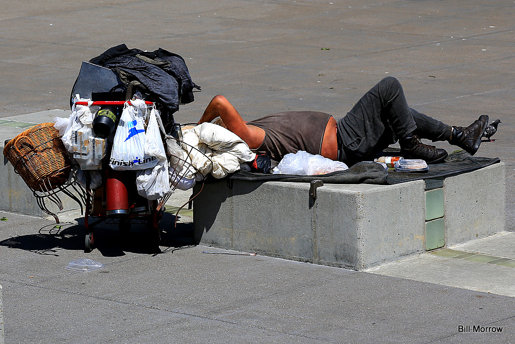 A man sleeping is sleeping on the street with his stuff under his head and some stuff hang on his cycle