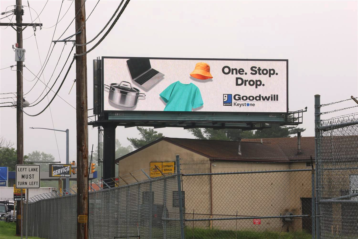 LED billboard advertising Goodwill with the logo, orange bucket hat, teal, t-shirt, laptop, and pot on the side of the road above a house.