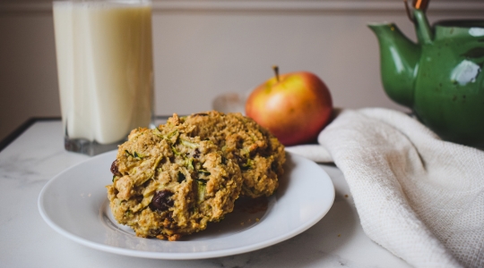 Deux biscuits aux courgettes et raisins secs sur une assiette blanche, accompagnés d'un verre de lait, d'une théière verte et d'une pomme.