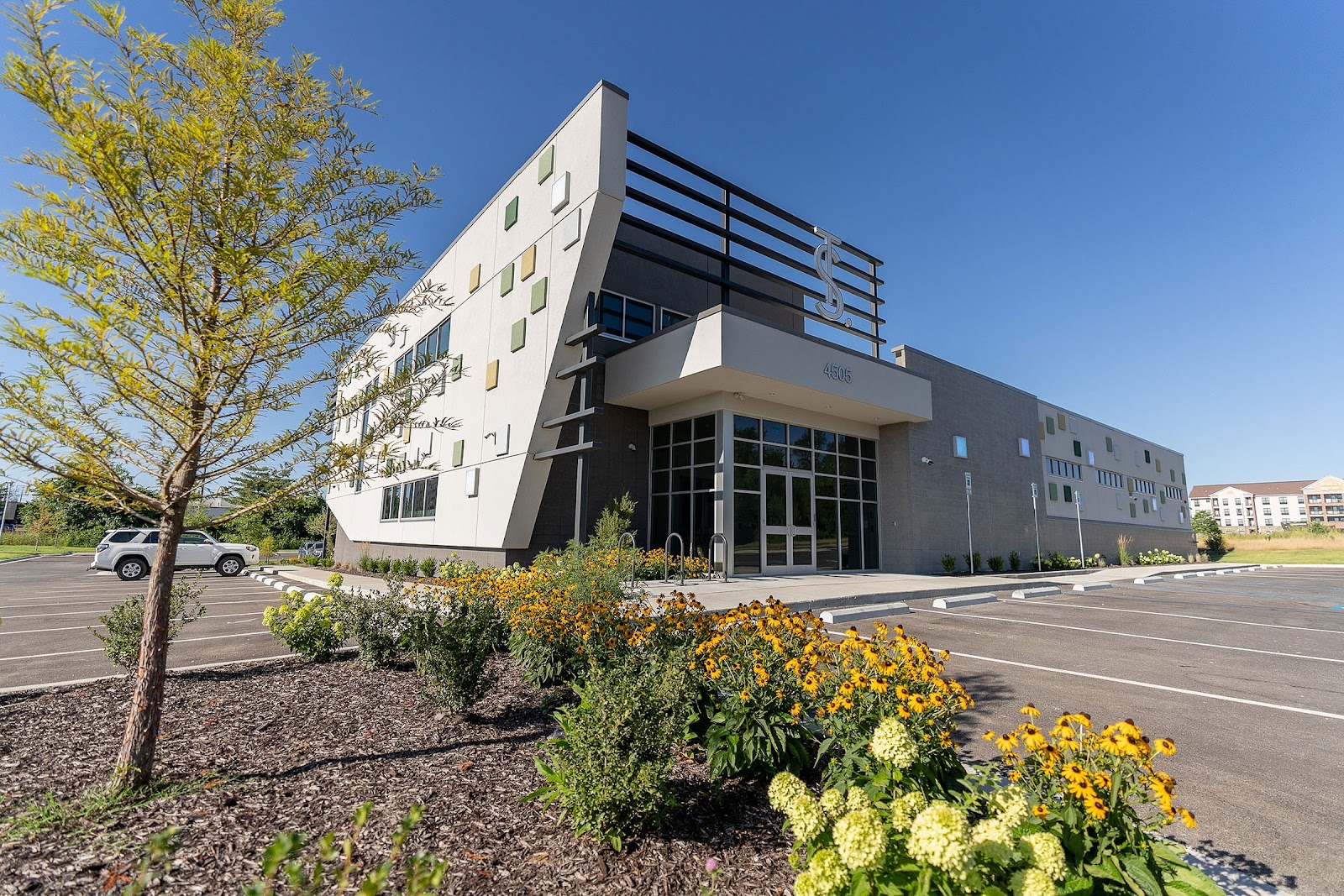 Exterior view of The Source's new state-of-the-art facility in Rogers, AR. The modern building features a striking white angular facade with colorful square accents, large windows, and The Source logo on top. The entrance is surrounded by a landscaped area with yellow flowers and young trees. A spacious parking lot is visible in the foreground. This multi-purpose facility houses the dispensary, grow operation, lab, kitchen, and Headwaters Provisions lifestyle store. 