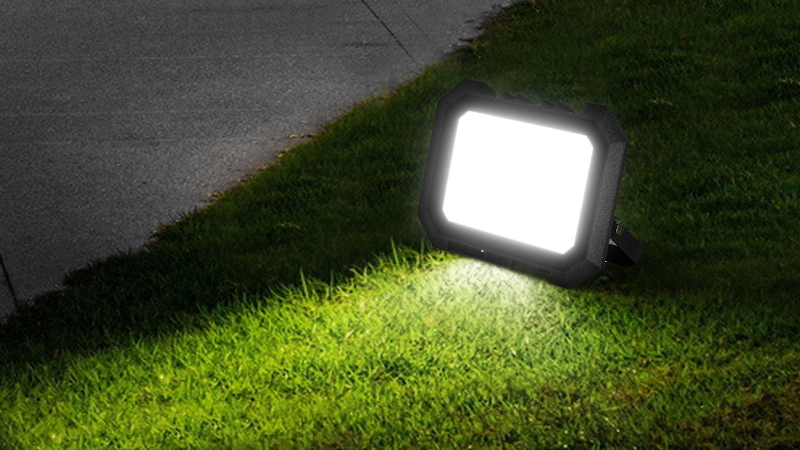 LED flood light placed on grass, casting bright illumination across lawn and sidewalk edge