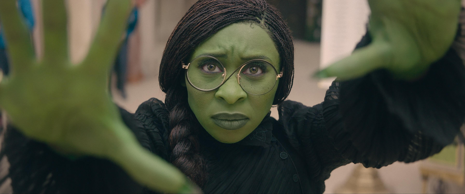 Elphaba (Cynthia Erivo) con piel verde y gafas, extendiendo las manos en un gesto dramático.