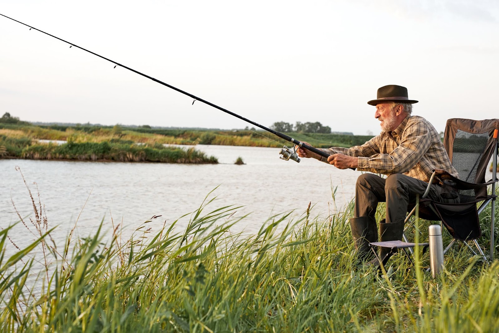 An older adult with a hat sits in a comfortable chair while fishing on the lake.