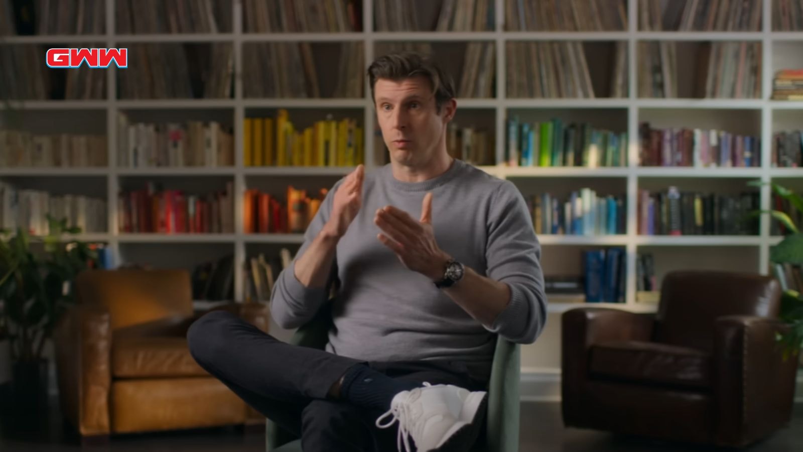 Matthew Reeve in a sweater gesturing, sitting in front of colorful bookshelves.