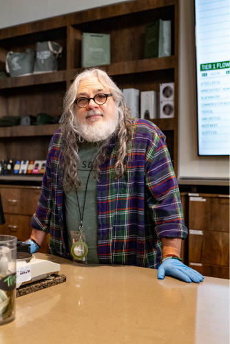 A long, silver-haired gentleman with glasses and a white beard stands at a budtending station at The Source cannabis dispensary in Rogers, Arkansas. 