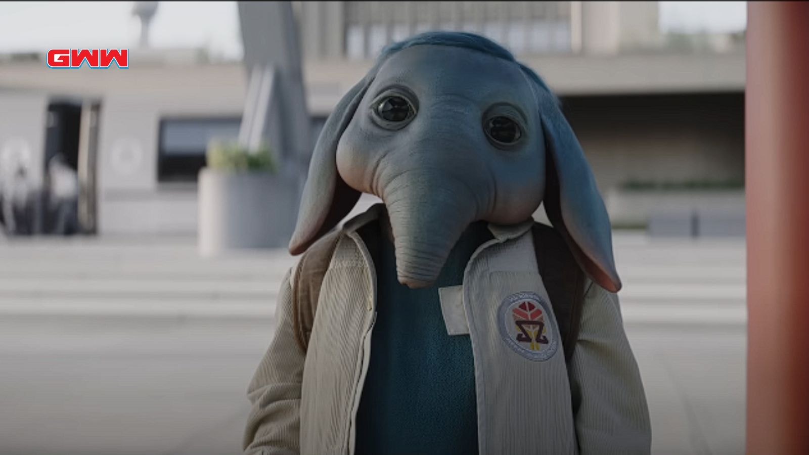 Robert Timothy Smith as an alien character with large ears wearing a jacket, looking into the camera.