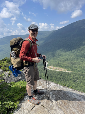 Man wearing a large backpack and smiling at the camera, standing on rock ledge overlooking a mountain valley.