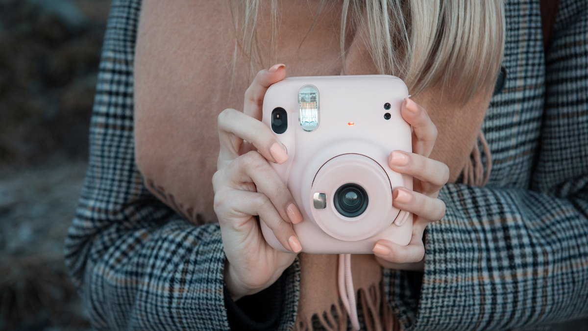 A person holding a pastel pink instant camera, ready to capture a moment, wrapped in cozy layers with a checkered coat and scarf.