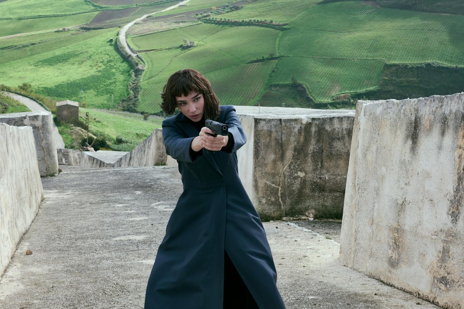 Escena de acción con una mujer apuntando una pistola, vestida con un abrigo largo oscuro. Está de pie en una estructura elevada con un paisaje verde montañoso de fondo, creando un fuerte contraste entre la acción y el entorno natural.